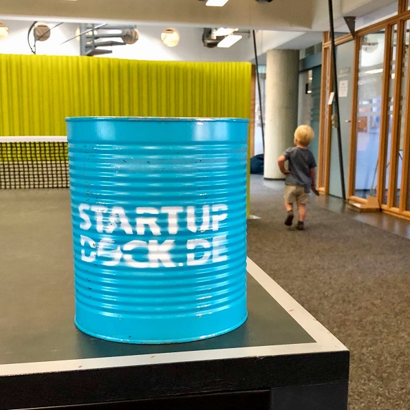 Foto aus dem Startup-Dock Hamburg mit einer Dose mit Beschriftung "Startup Dock" auf einer Tischtennisplatte mit Kind im Hintergrund